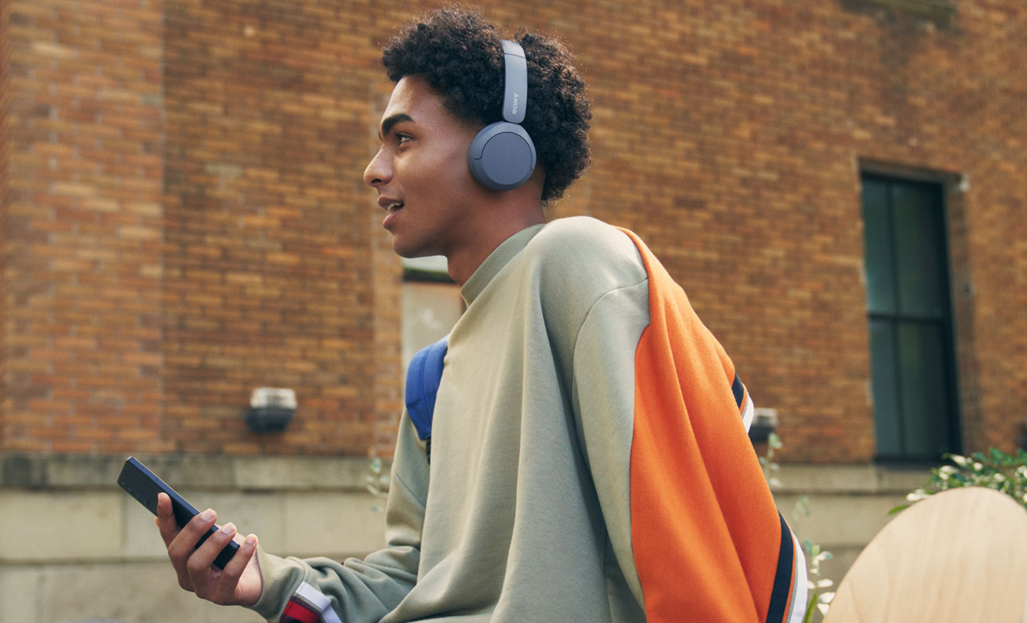 Sony WH-CH520 kulaklık takan, bir duvarın üzerinde oturan ve elinde cep telefonu tutan bir adamın, ön planda kaykayın yer aldığı görüntüsü