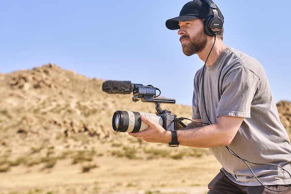 Çöl ortamında kulaklık takan ve büyük lens ile harici mikrofon takılı FX5 kamera kullanan bir videografın kullanım fotoğrafı.