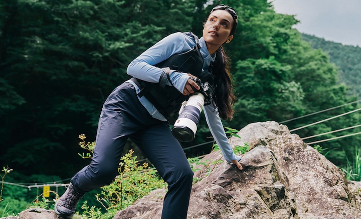 Bir elini nehir kenarındaki kayaya dayamış, süper telefoto lensli fotoğraf makinesi tutan fotoğrafçı
